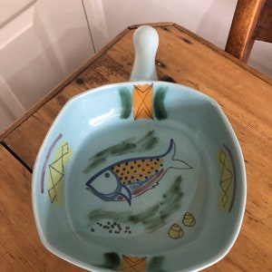 Puede incluir: Un plato para hornear de cerámica azul claro con asa. El plato tiene un diseño pintado a mano de un pez con detalles en amarillo, naranja y azul. También hay pequeños puntos amarillos y naranjas alrededor del pez.