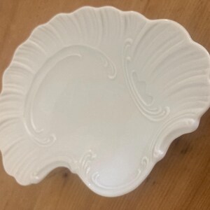 May include: A white ceramic dish shaped like a seashell, featuring a glossy finish and embossed, radiating ridges that mimic the natural texture of a scallop shell, with smooth, curved edges and a scalloped perimeter design.