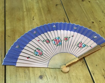 Puede incluir: Un abanico de mano con un diseño rosa claro y azul. El abanico presenta un borde azul y está decorado con motivos florales en rosa, azul y verde. El abanico está hecho de madera y papel y está abierto sobre una superficie de madera.
