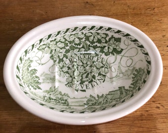 May include: An oval white ceramic soap dish featuring a green transferware pattern. The design includes a central floral motif with the text "CRABTREE & EVELYN" surrounded by a landscape scene with a horse-drawn cart and a rope-style border.