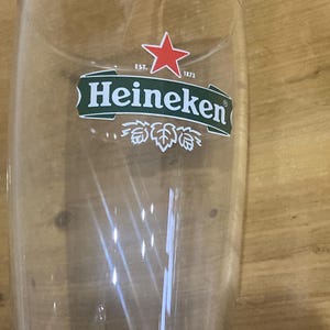 Op de afbeelding: Een helder, hoog bierglas met het Heineken-logo. Het logo toont een rode ster boven een groene banner met de merknaam en het jaar 1873. Het glas is leeg en lijkt van glas te zijn.