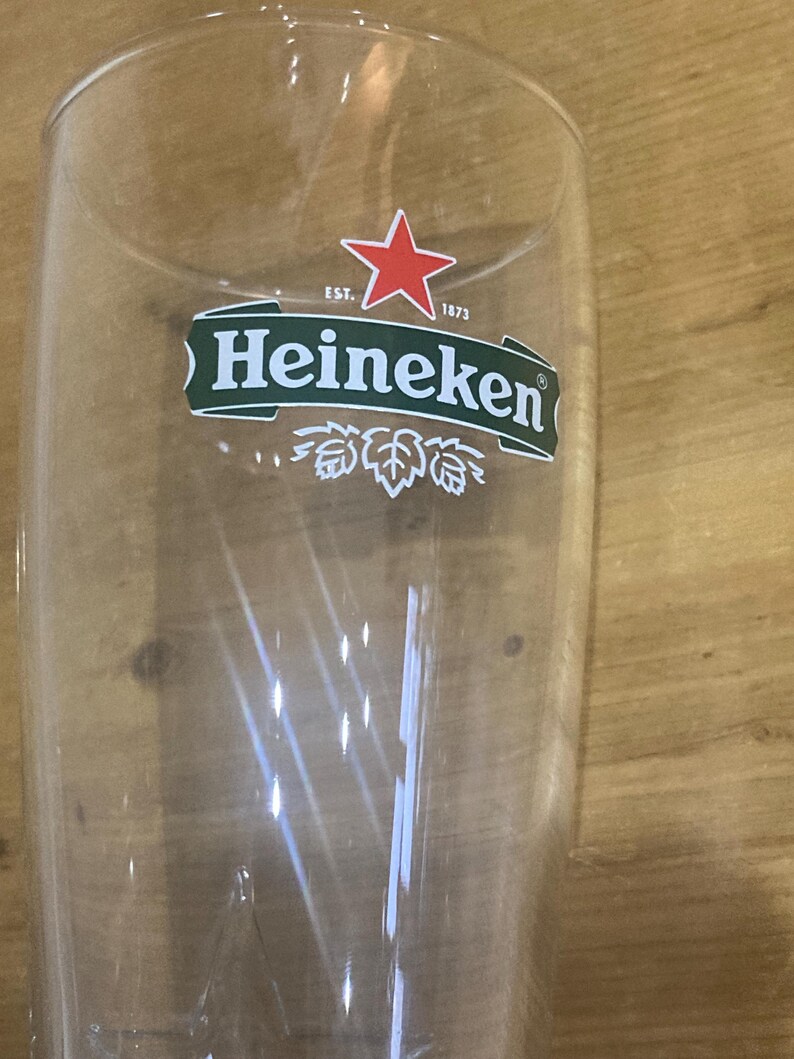 Op de afbeelding: Een helder, hoog bierglas met het Heineken-logo. Het logo toont een rode ster boven een groene banner met de merknaam en het jaar 1873. Het glas is leeg en lijkt van glas te zijn.