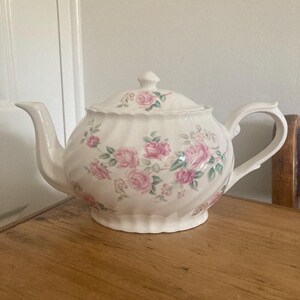 May include: A cream-coloured ceramic teapot featuring a fluted body and a delicate floral pattern of pink roses with green leaves. The teapot includes a matching lid with a small knob handle, a curved spout, and a sturdy handle on the side.