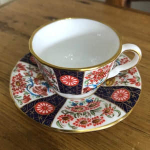Op de afbeelding: Een witte theekop en schotelset met een gouden rand. De kop en schotel hebben een bloemen- en geometrisch ontwerp in rood, blauw en wit. De theekop heeft een oor en staat op een bijpassende schotel.
