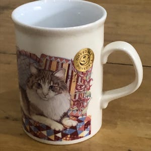 May include: A cream-coloured ceramic mug with a detailed illustration of a fluffy cat. The cat is depicted sitting on a patterned surface with floral and architectural elements. A gold sticker with the word DOVEDALE is on the side.