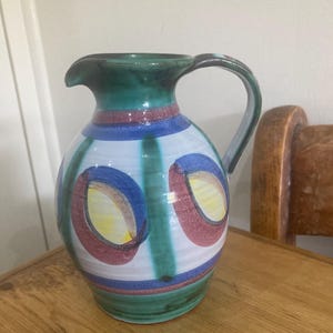 May include: A ceramic pitcher with a green handle and spout. The pitcher is decorated with blue, red, and yellow oval shapes and vertical green stripes on a white background. The base and rim are green.