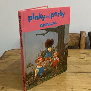 Op de afbeelding: Een vintage "Pinky and Perky Annual" boek met een roze kaft en blauwe letters. De kaft toont illustraties van de personages Pinky en Perky, en een vogel, in een boslandschap. Het boek staat op een houten tafel.