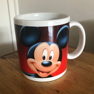 Puede incluir: Taza de cer&aacute;mica blanca con una imagen de Mickey Mouse en rojo, marr&oacute;n y azul.