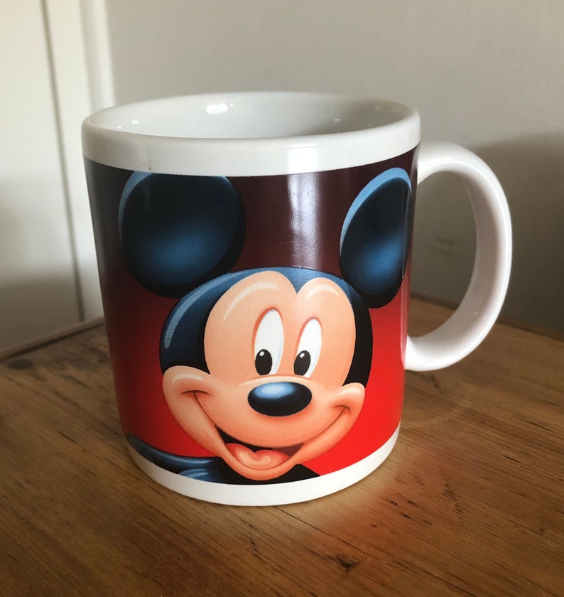 Puede incluir: Taza de cer&aacute;mica blanca con una imagen de Mickey Mouse en rojo, marr&oacute;n y azul.