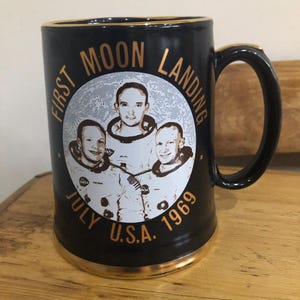 以下が含まれることがあります： 金色の縁取りと金色の文字で「First Moon Landing July U.S.A. 1969」と書かれた黒いセラミック製のマグカップ。マグカップには、1969年に月に降り立った3人の宇宙飛行士の円形の画像が描かれている。