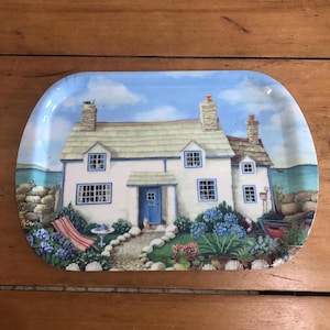 Könnte beinhalten: Dekorative Servierplatte mit Küstenhaus-Szene. Die Platte zeigt ein weißes Haus mit blauer Tür und Fenstern, umgeben von Blumen und einem kleinen roten Boot. Der Hintergrund zeigt einen blauen Himmel und das Meer.