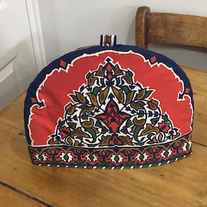 Op de afbeelding: Een gewatteerde theemuts met een levendig, sierlijk ontwerp. De theemuts heeft een rood middenpaneel met een gedetailleerd bloemmotief in wit, groen en bruin, omlijst door een marineblauwe rand. Bovenop zit een kleine lus.