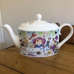 Könnte beinhalten: Weiße Keramik-Teekanne mit Deckel und Henkel. Die Teekanne ist mit einer farbenfrohen Illustration von Charakteren aus Alice im Wunderland verziert, darunter Alice, der Hutmacher und das weiße Kaninchen.