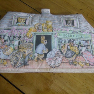 Può includere: Un giocattolo di peluche a forma di casa con un cartello che recita "Ginger & Pickles". La casa è decorata con una varietà di animali, tra cui conigli, gatti, galline e un'anatra. La casa è realizzata in un morbido tessuto rosa e ha una texture trapuntata.