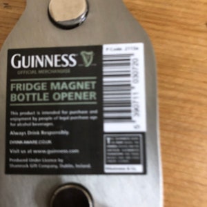 May include: A silver metal bottle opener shaped like a Guinness beer bottle with a black and white label that reads "GUINNESS OFFICIAL MERCHANDISE FRIDGE MAGNET BOTTLE OPENER". The label also includes the Guinness harp logo and a barcode. The bottle opener has two round magnets on the back.