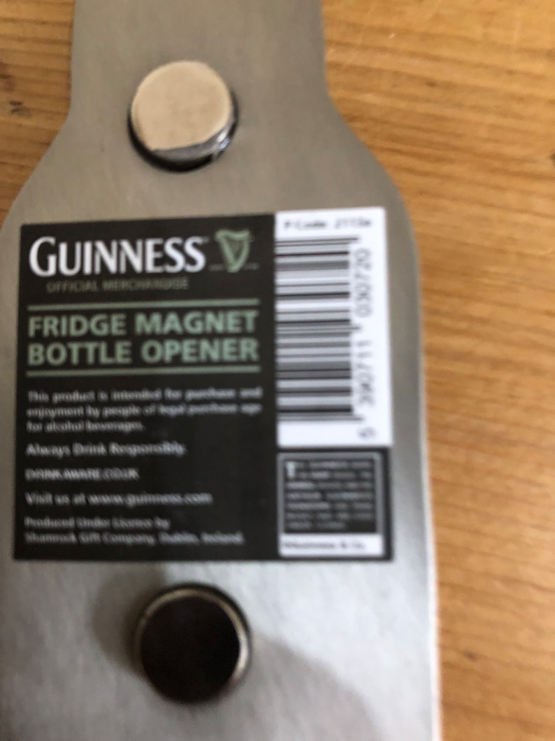 May include: A silver metal bottle opener shaped like a Guinness beer bottle with a black and white label that reads "GUINNESS OFFICIAL MERCHANDISE FRIDGE MAGNET BOTTLE OPENER". The label also includes the Guinness harp logo and a barcode. The bottle opener has two round magnets on the back.