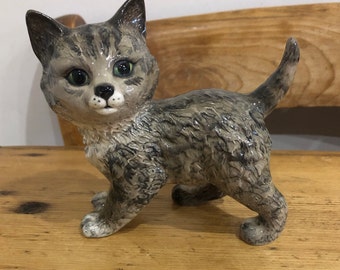 May include: A ceramic figurine of a grey tabby kitten with green eyes. The kitten is standing on all fours with its tail curled up behind it.
