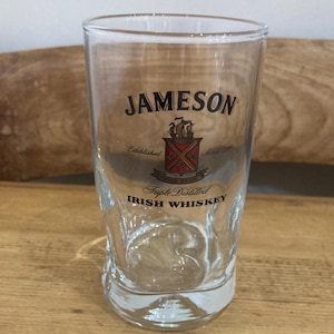 Può includere: Un bicchiere trasparente di Jameson Irish Whiskey con il nome del marchio e il logo stampati sul davanti. Il bicchiere ha una forma rettangolare con base arrotondata ed è appoggiato su una superficie di legno.