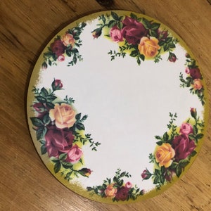 Könnte beinhalten: Ein runder weißer Kuchenteller mit goldfarbenem Rand und einem Blumendesign mit rosa, gelben und roten Rosen. Die Rosen sind von grünen Blättern umgeben.