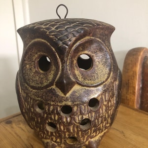 Op de afbeelding: Een bruine keramische uilvormige kandelaar met grote ooggaten en kleinere gaten op het lichaam. De uil heeft een getextureerd oppervlak en een metalen lus aan de bovenkant om op te hangen. De kleur is een mix van bruin en beige.