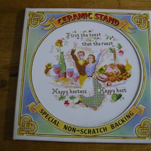 Puede incluir: Un soporte de cerámica blanco con un diseño bordado de colores que representa a una pareja brindando con copas de vino y un pollo asado. El soporte tiene un borde dorado y el texto "Ceramic Stand", "First the toast then the roast", "Happy hostess", "Happy host" y "Special Non-Scratch Backing".