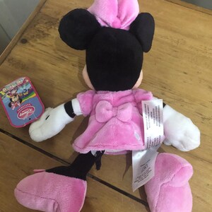 May include: Pink and black plush Minnie Mouse toy with a pink bow on her head. The toy is wearing a pink dress and has white gloves. The toy is sitting on a wooden surface.