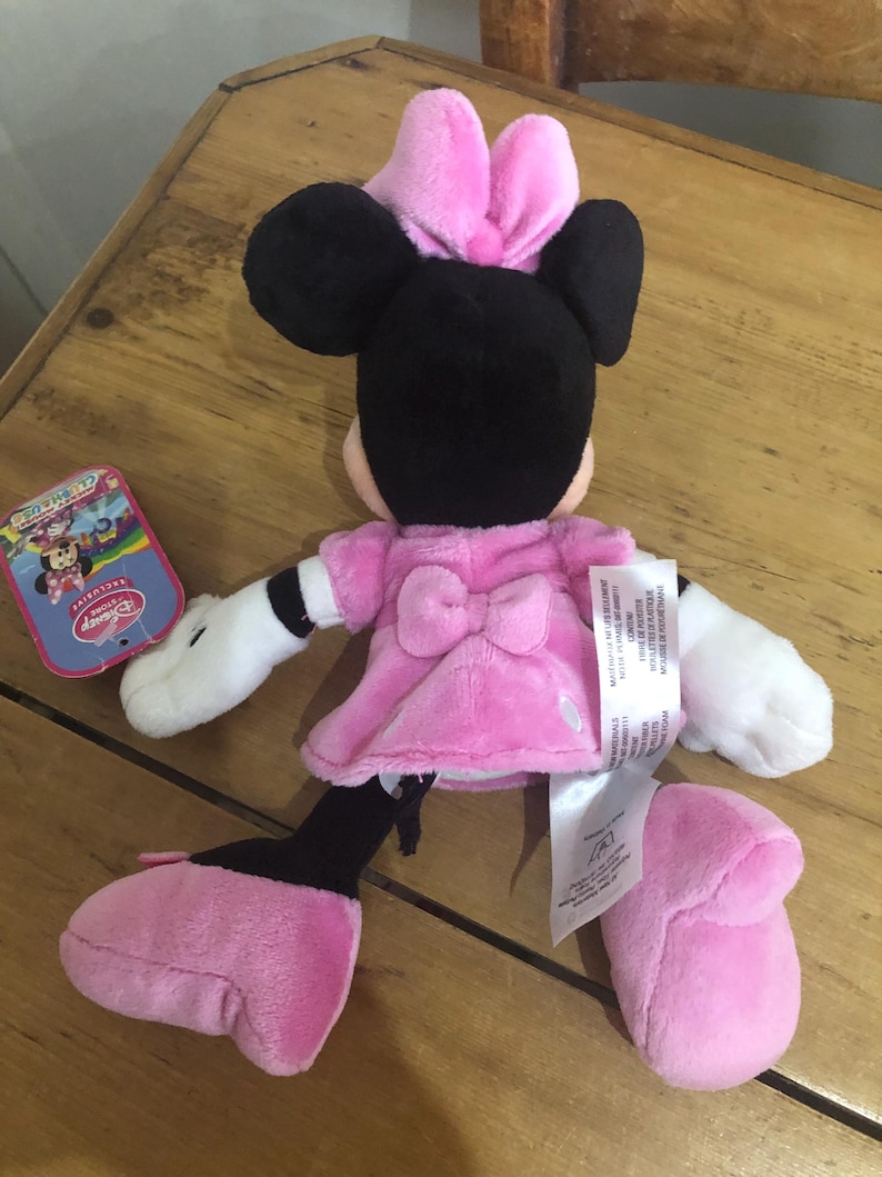 May include: Pink and black plush Minnie Mouse toy with a pink bow on her head. The toy is wearing a pink dress and has white gloves. The toy is sitting on a wooden surface.