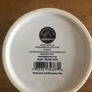 Puede incluir: Taza de cer&aacute;mica blanca con un dise&ntilde;o de Mickey Mouse negro. La parte inferior de la taza tiene una etiqueta negra con el texto "Everyone loves to get applause" y el logotipo de la empresa. La etiqueta tambi&eacute;n incluye el nombre de la empresa, la direcci&oacute;n, el sitio web y la informaci&oacute;n del producto.