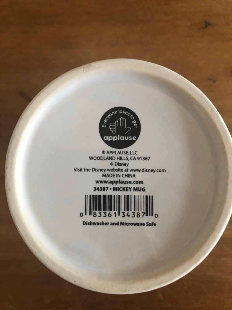 Puede incluir: Taza de cer&aacute;mica blanca con un dise&ntilde;o de Mickey Mouse negro. La parte inferior de la taza tiene una etiqueta negra con el texto "Everyone loves to get applause" y el logotipo de la empresa. La etiqueta tambi&eacute;n incluye el nombre de la empresa, la direcci&oacute;n, el sitio web y la informaci&oacute;n del producto.
