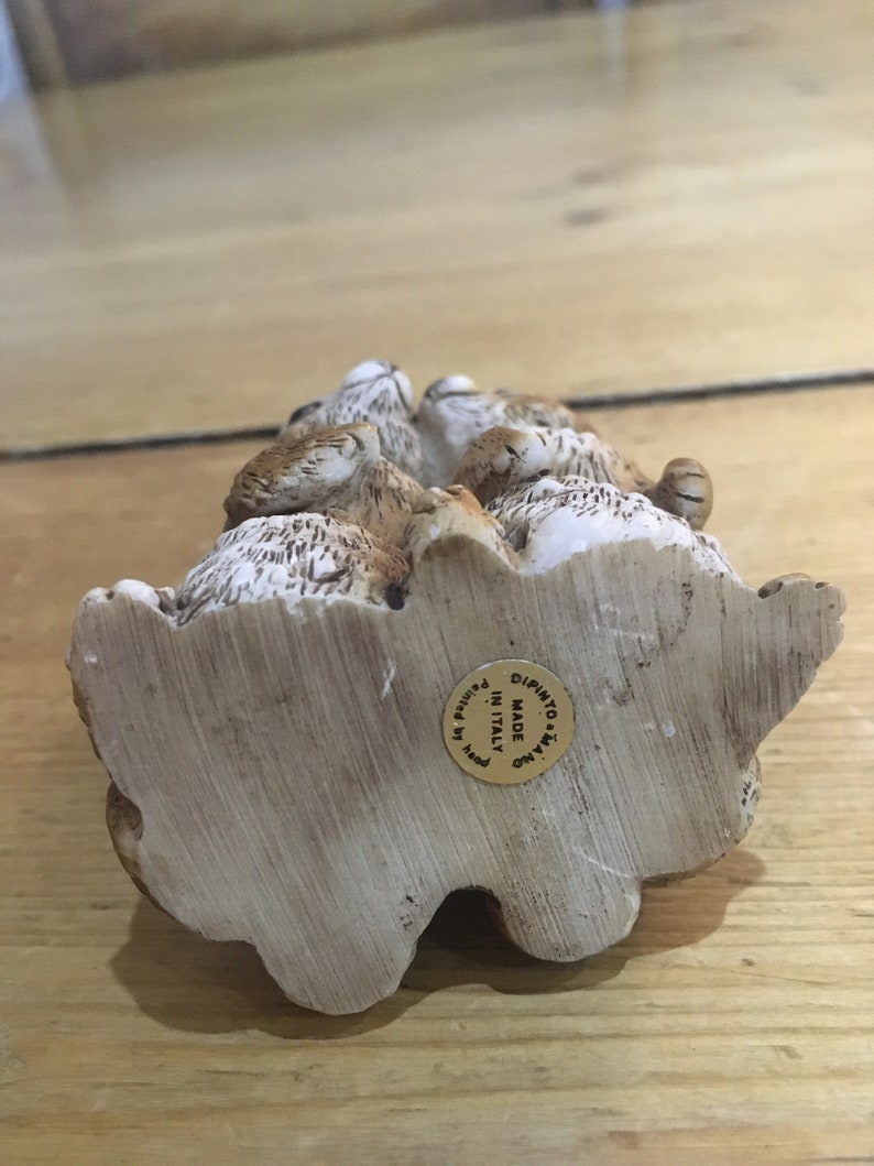 May include: A ceramic figurine of a group of rabbits, painted in shades of brown and white. The figurine is made in Italy and has a gold label on the bottom that reads "Made in Italy, Dipinto a Mano".