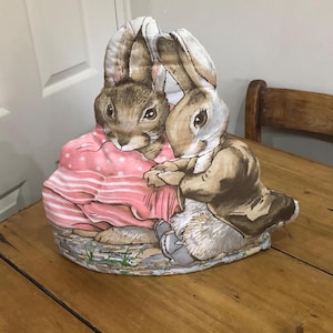 Puede incluir: Un cubrete de té decorativo con dos figuras de conejos. Uno de los conejos lleva un vestido rosa con lunares blancos, el otro una chaqueta marrón. El cubrete está sobre una mesa de madera.