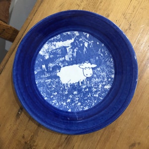 May include: A blue and white ceramic plate with a white sheep illustration in the center. The plate has a blue rim and a white background with a blue floral pattern.