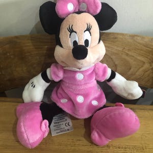 May include: Pink Minnie Mouse plush toy with white polka dots on her dress and bow. The toy has black eyes and a black nose. The toy is sitting on a wooden surface.