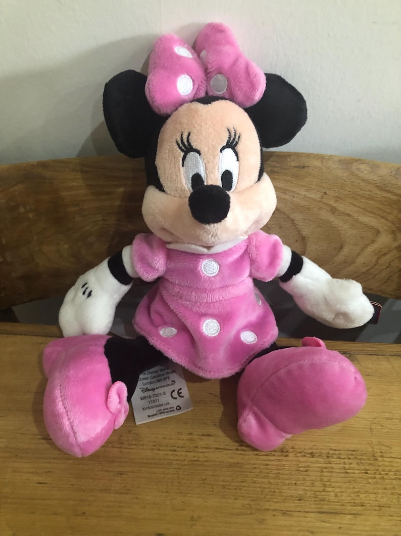 May include: Pink Minnie Mouse plush toy with white polka dots on her dress and bow. The toy has black eyes and a black nose. The toy is sitting on a wooden surface.