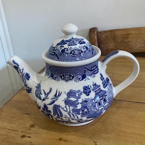 Può includere: Una teiera in ceramica bianca con un motivo blu Willow. La teiera presenta un beccuccio curvo, un corpo arrotondato e un manico. Il coperchio ha un piccolo pomello. Il design include pagode, uccelli ed elementi floreali.