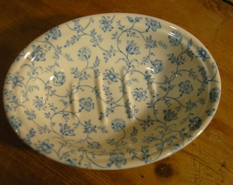May include: Oval-shaped ceramic soap dish with a white background and a blue floral pattern. The dish has a slightly raised rim and three drainage slots. The floral design features delicate blue flowers and vines.