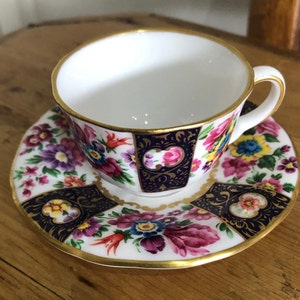 Op de afbeelding: Een wit theekopje en schotel met een bloemenpatroon in tinten roze, paars, geel en blauw. Het kopje en de schotel hebben een gouden rand en een donkerblauwe achtergrond.