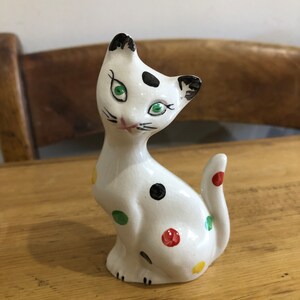 Puede incluir: Una figurita de cerámica blanca de un gato con lunares negros, rojos, verdes y amarillos. El gato tiene ojos verdes y marcas negras en la cara y las orejas.