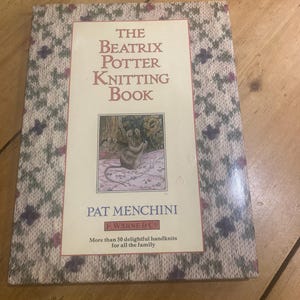Puede incluir: Un libro de tapa dura titulado "The Beatrix Potter Knitting Book" de Pat Menchini. La portada presenta un borde con un patrón de punto en tonos grises, verdes y morados. El título del libro está en letras grandes y la portada también incluye una ilustración.