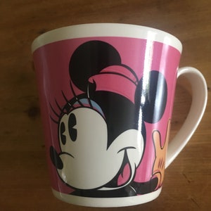 Peut inclure: Une tasse en c&eacute;ramique blanche avec un fond rose et un motif de Minnie Mouse. Le motif comprend le visage de Minnie, des oreilles noires et un n&oelig;ud rose. La tasse a une anse et un bord blancs.