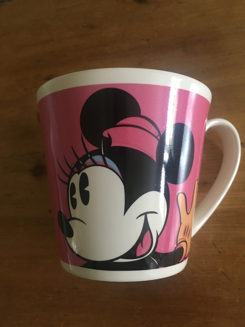 Peut inclure: Une tasse en c&eacute;ramique blanche avec un fond rose et un motif de Minnie Mouse. Le motif comprend le visage de Minnie, des oreilles noires et un n&oelig;ud rose. La tasse a une anse et un bord blancs.