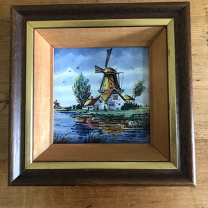 Könnte beinhalten: Ein gerahmtes Fliesenbild mit einem holländischen Windrad, blauem Himmel und grünem Gras. Das Windrad ist gelb und weiß mit einem roten Dach. Im Vordergrund befinden sich Bäume und ein kleines Haus.