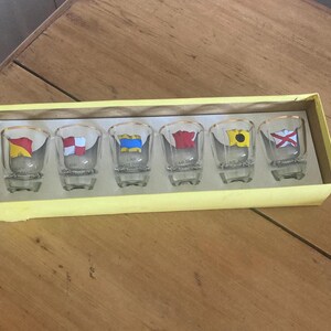 May include: A set of six clear glass shot glasses with gold rims, each featuring a unique colorful nautical signal flag graphic and text printed on the front, housed in a rectangular yellow cardboard display box with a white interior tray.