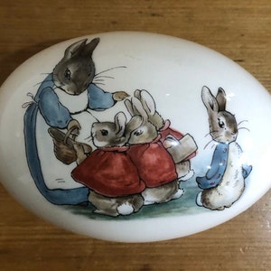 Puede incluir: Una caja de cerámica blanca en forma de huevo con una imagen pintada de Peter Rabbit y su familia. Los conejos llevan ropa colorida y están de pie en un campo.