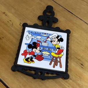 May include: Black metal trivet with a square ceramic tile featuring Mickey and Minnie Mouse cooking. The tile shows a blue stove, a pie in the oven, and the characters in chef attire. The trivet has a decorative handle.
