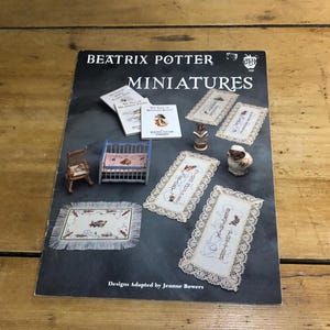 May include: A book cover featuring Beatrix Potter Miniatures. The cover shows a variety of miniature items, including a cot, a rocking chair, and several small rugs. The book cover is black with white text and a small red strawberry logo.
