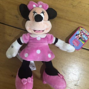 May include: Pink Minnie Mouse plush toy wearing a pink dress with white polka dots and a pink bow. The toy has black eyes and a black nose. The toy is holding a tag that says "Disney Junior".