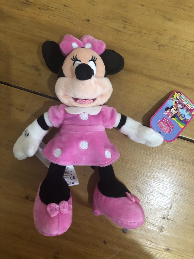 May include: Pink Minnie Mouse plush toy wearing a pink dress with white polka dots and a pink bow. The toy has black eyes and a black nose. The toy is holding a tag that says "Disney Junior".