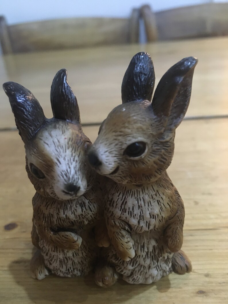 May include: Two brown and white ceramic rabbit figurines standing side by side. The rabbits have black ears and brown fur with white markings.