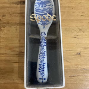 May include: A white ceramic spoon with blue floral and landscape designs, presented in a box. The spoon's handle features blue floral patterns, while the bowl displays a blue landscape. The word "Spode" is printed on the spoon.